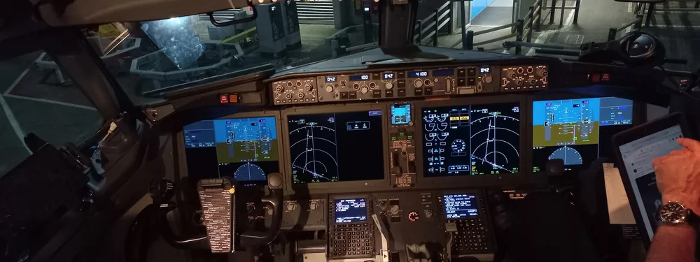 Boeing 737 MAX flight deck with large display screens, a lit overhead panel, and a crew member’s arm at the right edge
